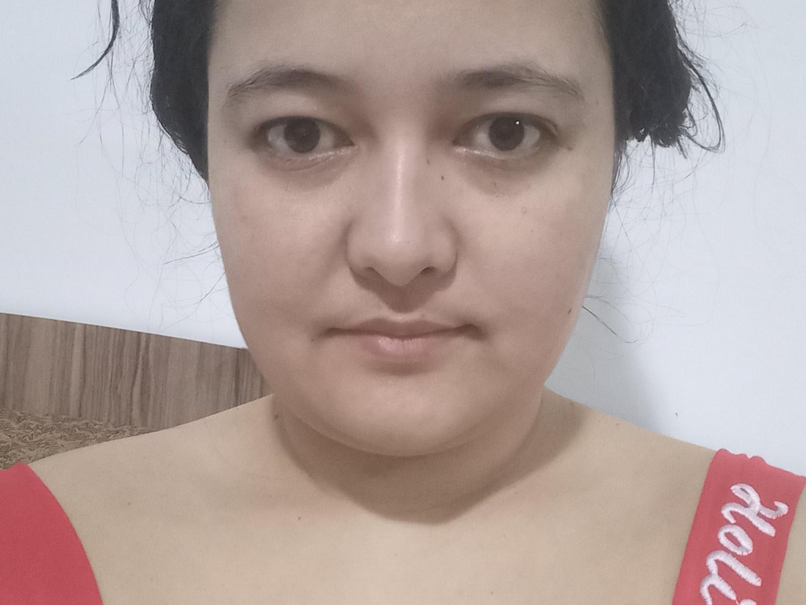 MollyJanette's face, showing a neutral expression with brown eyes, light skin, and black hair in a messy updo. She's wearing a red top with white text.