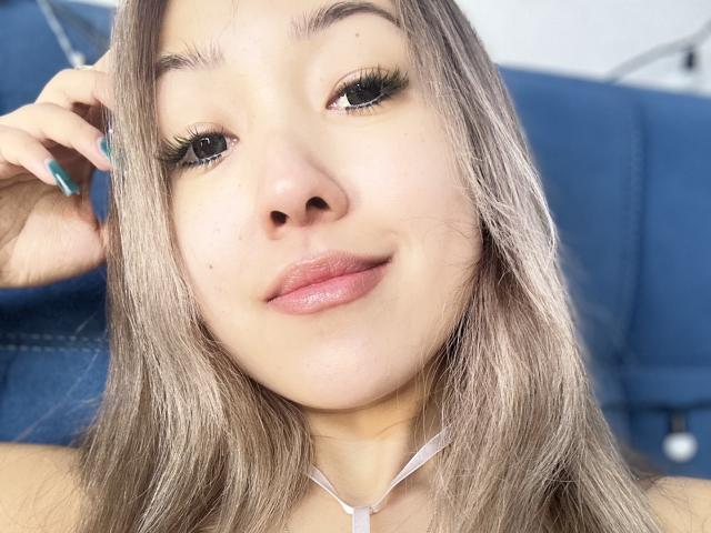 RenyLime's face, showing a slight smile with pink lips, long grayish-brown hair, and teal nail polish, wearing a white necklace, with blue upholstery in the background.