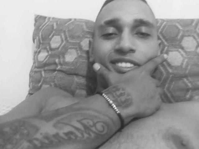 Black and white photo of BrunetteX69 with short hair, smiling, holding his chin with a tattooed hand, showing a bracelet on his wrist. His nose is slightly upturned.