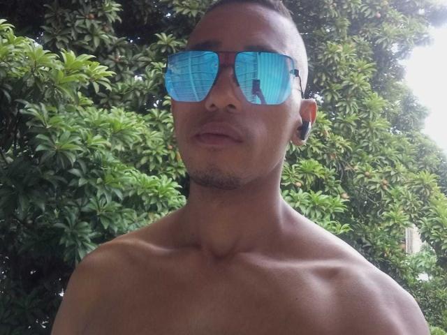 BrunetteX69 with blue reflective sunglasses, short brown hair, and a bare, muscular, tan chest. His face has a slight mustache and he's looking slightly to the right.