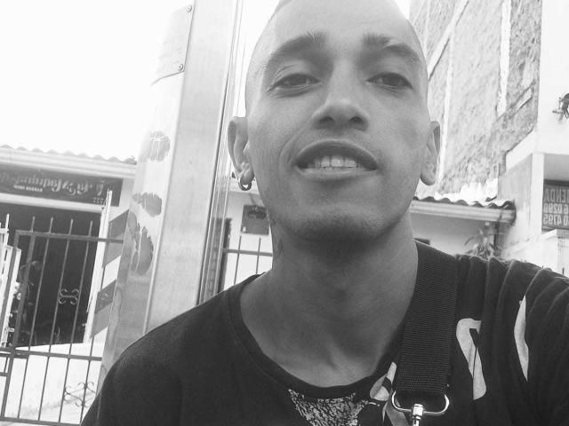 Black and white photo of BrunetteX69 with bald head, small earrings, dark shirt, necklace, and visible teeth, standing outside with a metal fence in the background.
