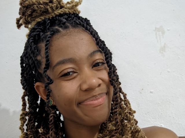 AmahI has dark brown skin, black and blonde twisted braids, a top bun, green earrings, and a subtle smile with a slightly crooked nose.