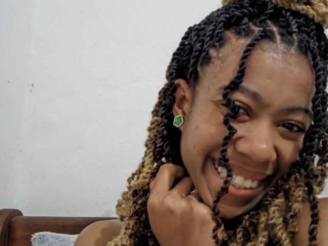 AmahI has dark brown skin, black and blonde braids, green earrings, and a happy smile, holding her chin with her hand. She's showing off her teeth.