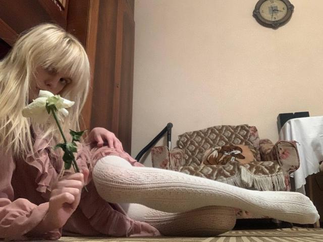 Lialit with blonde hair, pink dress, and white knee socks, holding a white rose, sitting on carpet, leg raised, looking shy. Clock on beige wall, patterned chair.