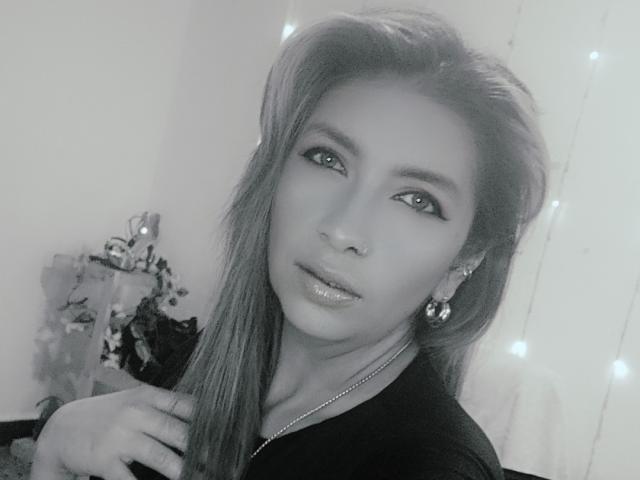MichelleDumont with long hair, shiny lips, and dark eyeliner, wearing earrings and a necklace, touching her hair, in a black top, with soft fairy lights in the background.