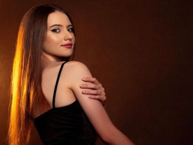 NatashaAimee with long brown hair, black strap dress, and a hand on her bare shoulder, looking over her shoulder with a slight smile, warm light on her face.
