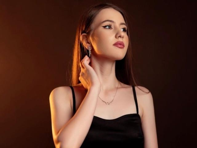 NatashaAimee has long brown hair, wearing a black sleeveless top, with her hand near her ear, looking to the side, showing her tits slightly in the dim light.