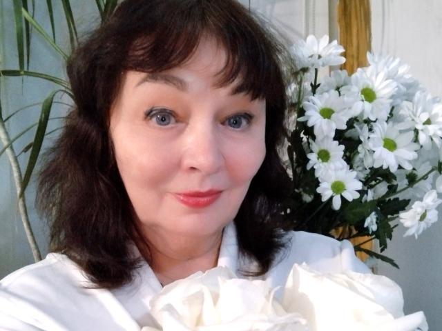 MilaMils with medium brown wavy hair, blue eyes, red lipstick, wearing a white shirt, holding white daisies, smiling softly.