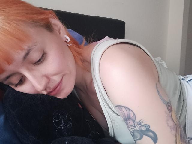 SaraleePoison with orange hair, closed eyes, smiling, wearing a green tank top, showing a tattoo on her right arm, lying on a dark blanket.