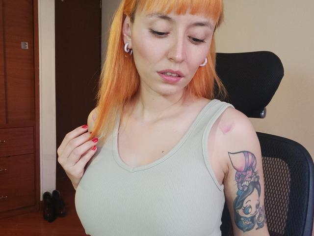 SaraleePoison with orange hair, white tank top, and tattoos, has small boobs, red nail polish, and a bruise on her right shoulder, sitting in a black office chair.