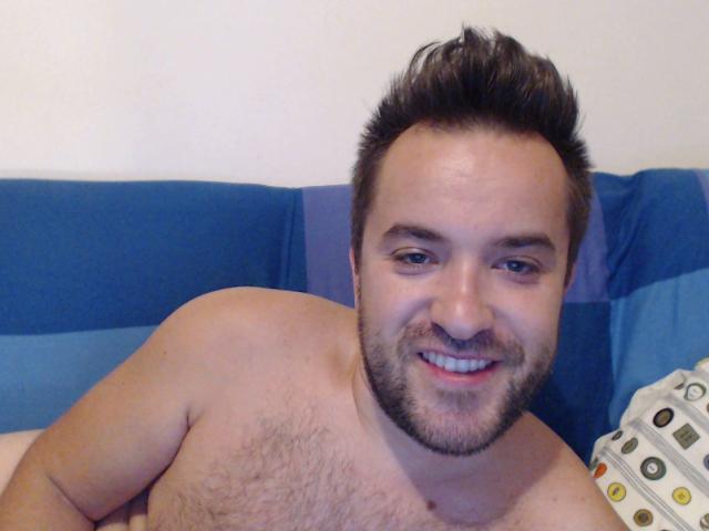 GabrielNastyGuy sem camisa, com um sorriso feliz, cabelo castanho espetado, barba e peito peludo, sentado em um sofá azul, segurando um controle remoto. Seus olhos azuis estão ligeiramente semicerrados..