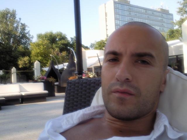 GabrielNastyGuy, bald with a slight stubble, white shirt, medium build, tan skin, relaxed, leaning back, outdoor setting, sunlight, blue sky, white building in background, trees, wicker chair.