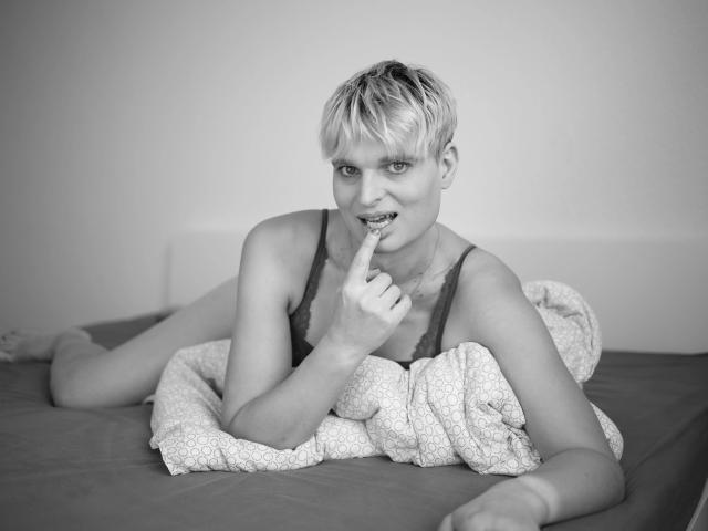 LolaAnders, une femme attrayante aux cheveux blonds courts, allongée sur un lit, portant un soutien-gorge en dentelle, un doigt dans la bouche, souriante, couverte d'une couverture à motifs, sur une photo en noir et blanc..