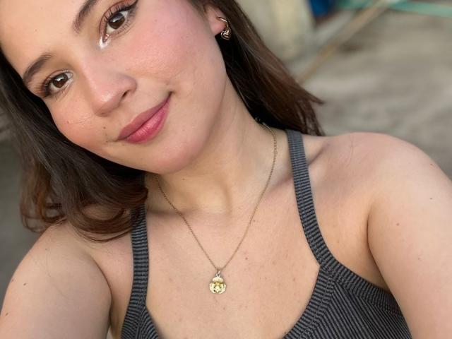 CelesteDash, avec ses yeux marron, ses lèvres roses et son petit nez, portant un débardeur gris et un collier en or avec un pendentif en forme de cœur, souriant à l'appareil photo..