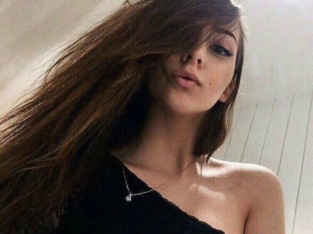 MariaKissYou with long brown hair, black one-shoulder top, and subtle makeup, wearing a small necklace, looking slightly to the side with a relaxed expression.