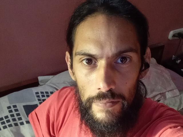 Morochoo69 ha un viso marrone, lunghi capelli neri raccolti in una coda di cavallo, occhi marroni e una camicia rossa con una barba scura. È sdraiato a letto con un cuscino..