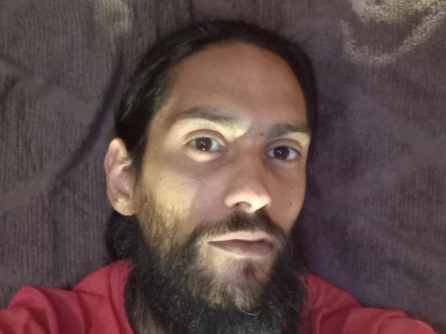 Morochoo69 a une peau marron moyen, une barbe noire et de longs cheveux noirs. Il porte une chemise rouge, est allongé et a un léger sourire. Ses yeux sont tournés vers le haut..