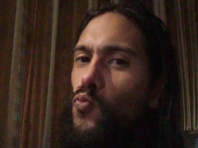 Morochoo69 a une barbe foncée, des lèvres fines et de longs cheveux bruns. Il regarde sur le côté avec une expression légèrement sérieuse, dans un environnement peu éclairé..