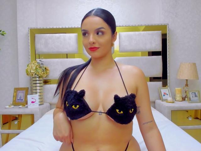 NovaDixon met een zwart bikin met kattenoren, die haar borsten laat zien, met rode lipstick, lang zwart haar in een staart, een tatoeage op haar rechterarm, staand in een slaapkamer..