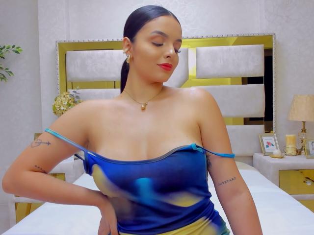 NovaDixon with dark hair in a ponytail, wearing a blue and yellow off-shoulder dress, red lipstick, gold earrings, and a necklace, showing tattoos on her arm.