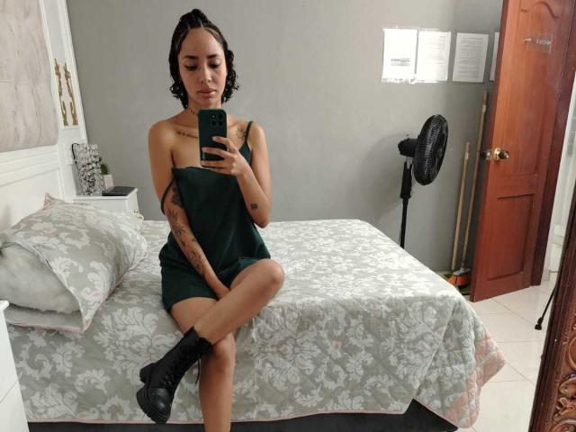 SilvanaXOwen with short curly hair, green strapless dress, black boots, tattoos on arm, sitting on bed, holding phone, taking mirror selfie, showing bare tits and ass.