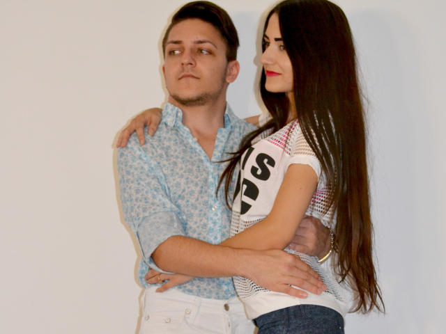 HotCoupleAnal: Hombre con cabello castaño corto, con una camisa azul estampada, con los brazos alrededor de una chica de cabello castaño largo, vestida con una blusa blanca y rosa, mirando hacia un lado. Las manos de ambos están en los glúteos del otro..