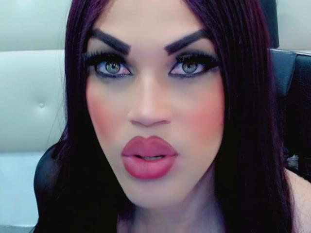 AlissonSaen's face, with full pink lips, thick dark eyebrows, green eyes, and long purple hair. She's looking directly at the camera with a slightly open mouth.
