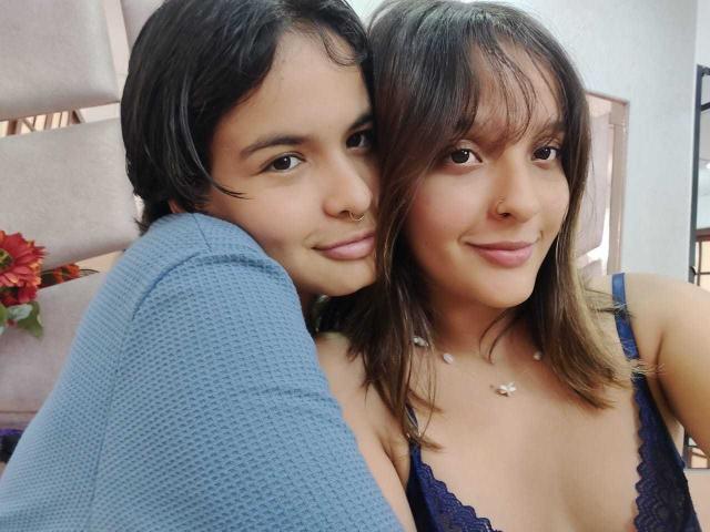 AnneAndSophia with short dark hair and nose ring, blue sweater, hugging Sophia with brown hair, blue lace bra, smiling, both showing small tits, in selfie.
