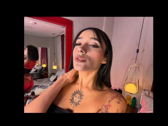 SalomeMille with black hair, sun tattoo on chest, floral tattoo on shoulder, holding neck, red lipstick, in bedroom with red mirror, hanging lamp, and yellow light.