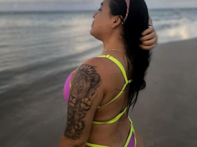 Beatrixxzz with long black hair, wearing a neon yellow and purple bikini, showing a large tattoo on her right arm, standing by the ocean. Her hand touches her head.