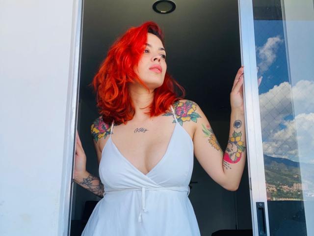 CandyDelirio mit leuchtend roten Haaren, Tätowierungen an Armen und Brust, trägt ein weißes Kleid mit tiefem Ausschnitt, steht in einem Türrahmen, blickt nach oben, Hände an der Tür..