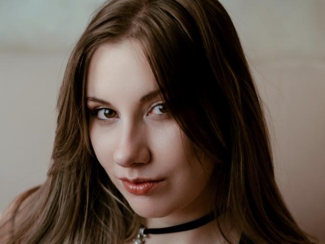 RinDragonesse has long brown hair, a black choker, and a subtle smile with red lips, looking directly at the camera with brown eyes. Her skin is pale and smooth.