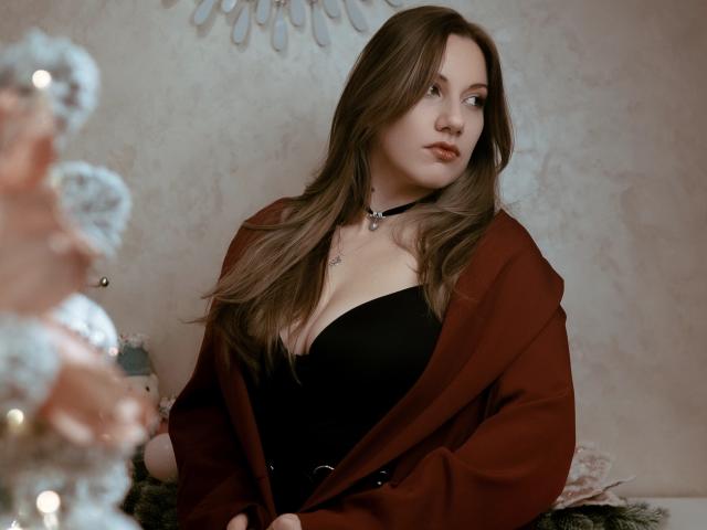 RinDragonesse with long brown hair, wearing a black strapless top, red coat, black choker, and necklace, looking to the side with a slight smile. Boobs are visible.