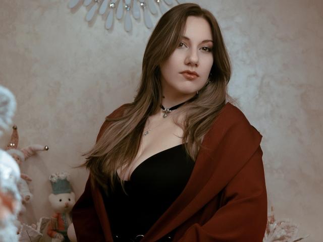 RinDragonesse ha un seno di media grandezza, indossa un top nero senza spalline, un cappotto rosso, ha lunghi capelli castani e una collana, e guarda leggermente di lato in una luce soffusa..