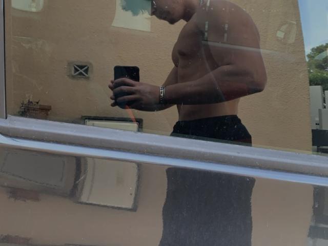 MagicalMike, torse nu, musclé, peau foncée, debout, pantalon noir, tenant un téléphone, reflet dans la fenêtre, montre argentée, cheveux bleus, léger sourire, regardant vers le bas, journée..