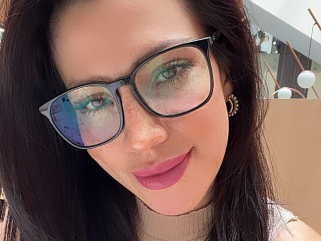 Liliysweet with dark brown hair, freckles, and pink lipstick, wearing black cat-eye glasses and gold earrings, smiling softly. Her reflection in the glasses is visible.