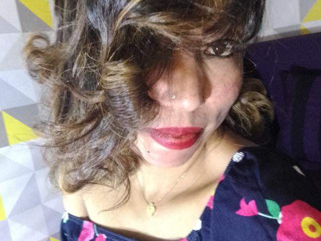 AnniaTattoo with curly brown hair, red lipstick, nose piercing, and floral dress, wearing a gold necklace, looking at camera. Her tits are not visible.