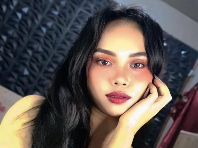 Tsmaja69 has dark, wavy hair, pinkish-orange eyeshadow, and red lipstick. She's touching her cheek with her right hand, looking directly at the camera with a slight smile.