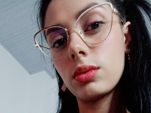 AnaisHilton has glasses on, dark hair, red lipstick, and is looking into the camera with a slightly pouty expression. Her ass is not visible.