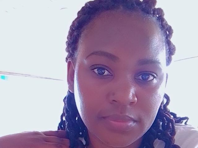 SweetFountaine has dark brown, curly hair, medium brown skin, and blue eyes. She's looking slightly down with a subtle smile, showing her nose and lips. Her hand's near her neck.