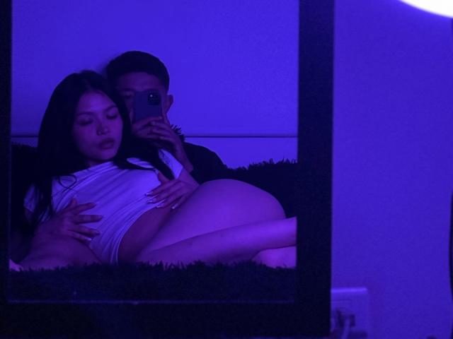 SarayDiego with black hair, white top, and bare ass, lying on a dark bed, held by a guy with a cap, taking a mirror selfie in purple light.
