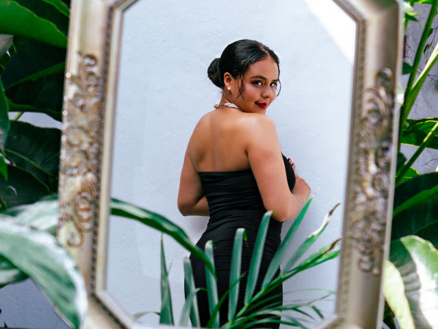 Arielle Wilde mit dunklem Haar zu einem Dutt gebunden, trägt ein schwarzes Kleid ohne Träger und blickt über ihre Schulter in einen Spiegel mit aufwendigen Rahmen, umgeben von grünen Pflanzen..