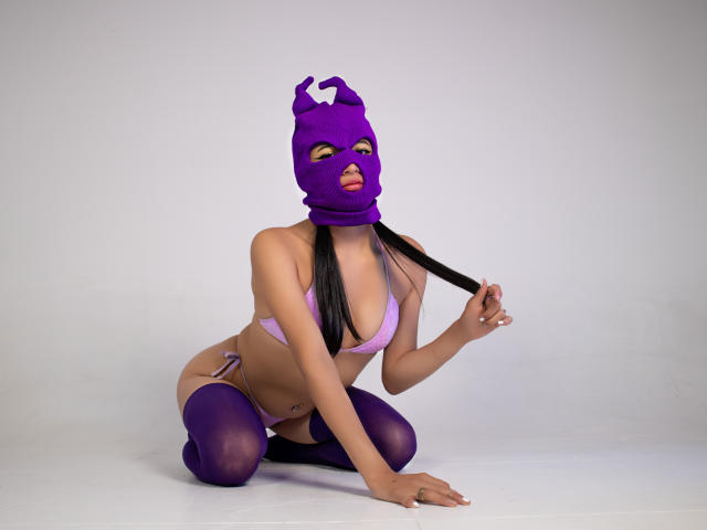 NaiaSanders kneels in purple lingerie, mask, and thigh-highs, playing with her black pigtails, exposing her tan skin and small, perky tits. Her tongue slightly peeks through the mask.