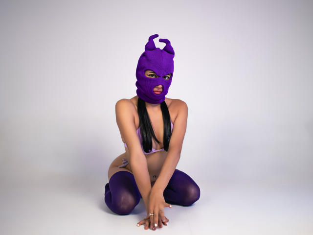 NaiaSanders, wearing a purple mask, black bra, and purple thigh-highs, kneels with her tits exposed, long black hair, and a tattoo on her right side.