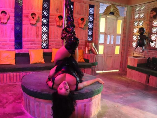 AlexaPaulfer is upside down on a circular platform, wearing black latex and boots, with her tits exposed and her ass in the air, in a sexy, suspended pose.