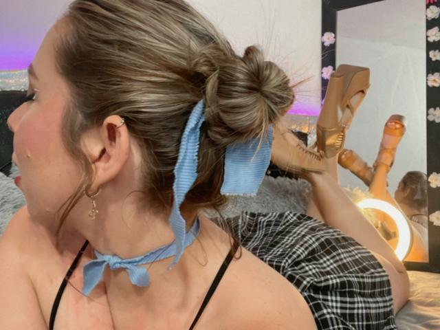 JennyLawry with her hair in a bun, wearing a blue ribbon and choker, black strap top, plaid skirt, tan high heels, lying on bed, kissing mirror.