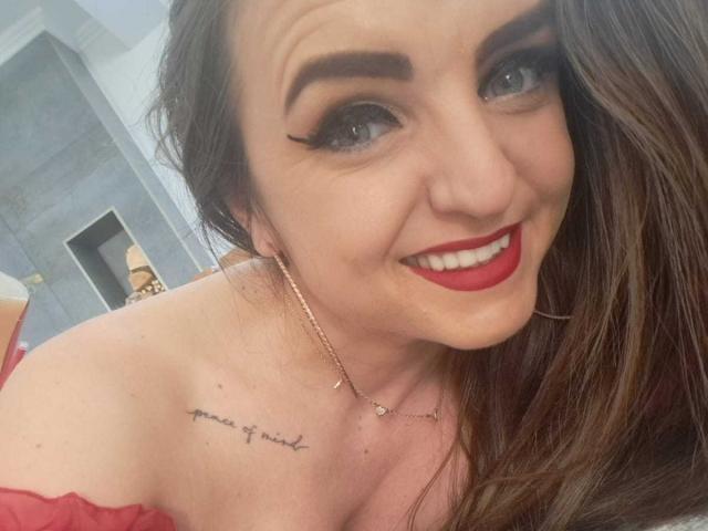 WildBlondy69 with red lipstick, brown hair, and a tattoo on her shoulder that reads "my heart is" smiling at the camera, showing her tits in a red off-shoulder top.