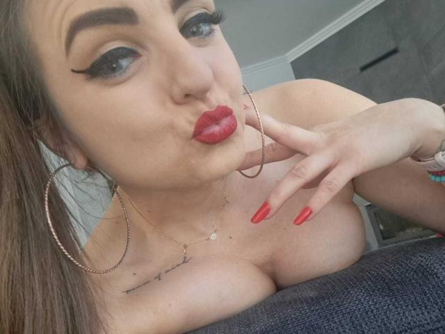WildBlondy69 with red lipstick, long red nails, and large hoop earrings, pouting and touching her chin. Her tits are visible, and she has a tattoo under her right breast.
