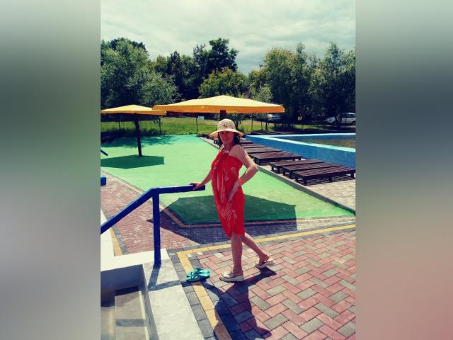 BellisimaAh wears a red dress and sunhat, standing on a brick staircase with blue railing, holding a green water bottle, next to yellow umbrellas and a green pool.
