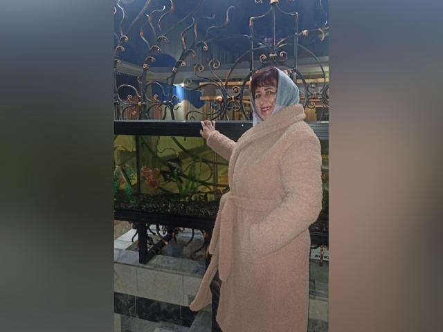BellisimaAh with short brown hair, blue hijab, and beige fluffy coat, smiling, pointing with right hand, standing next to ornate metal and glass wall.